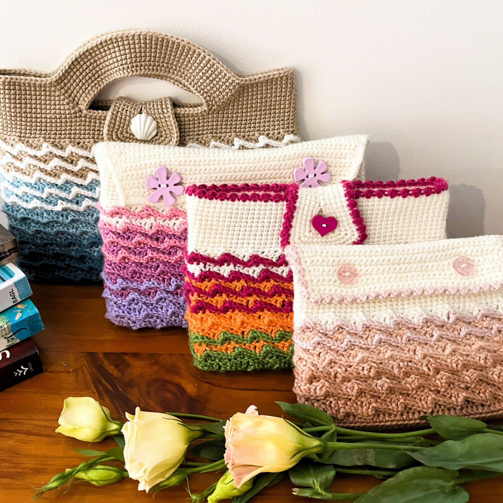 Collection of crocheted bags and pouches on a wooden surface with books and flowers.