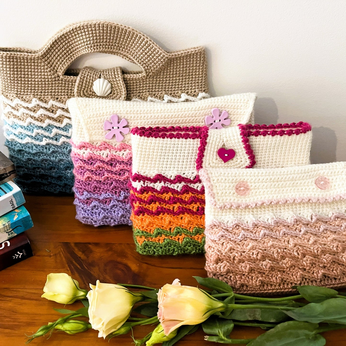 Collection of crocheted bags and pouches on a wooden surface with books and flowers.
