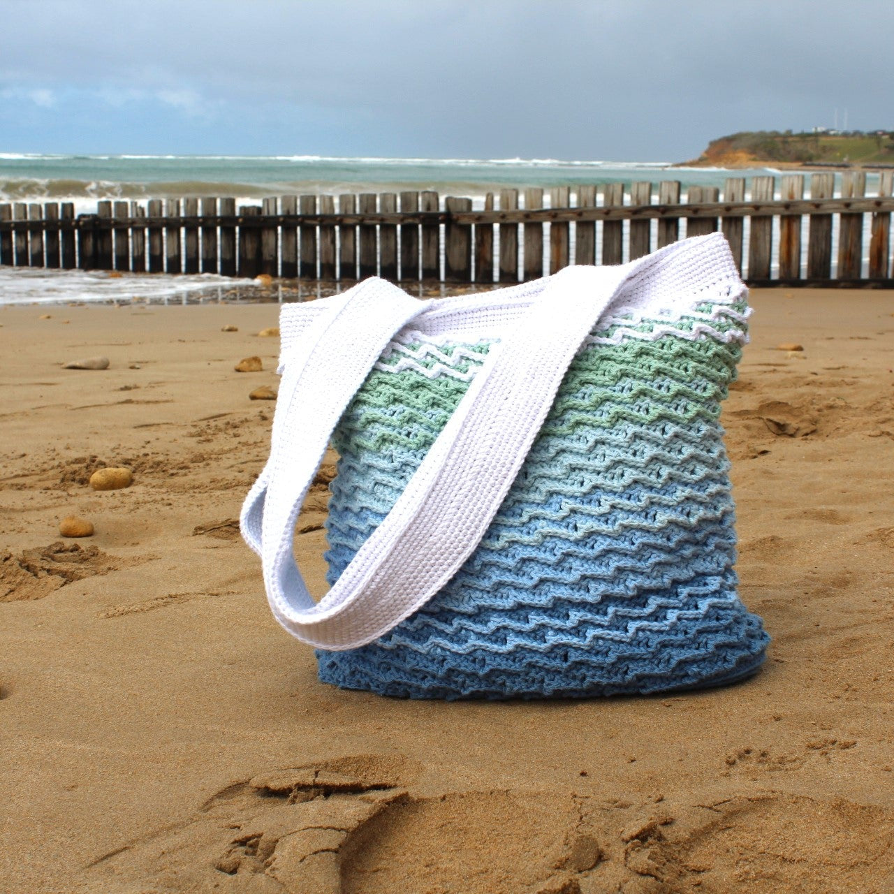 Blue and green woven bag on a sandy beach with ocean and pier in the background