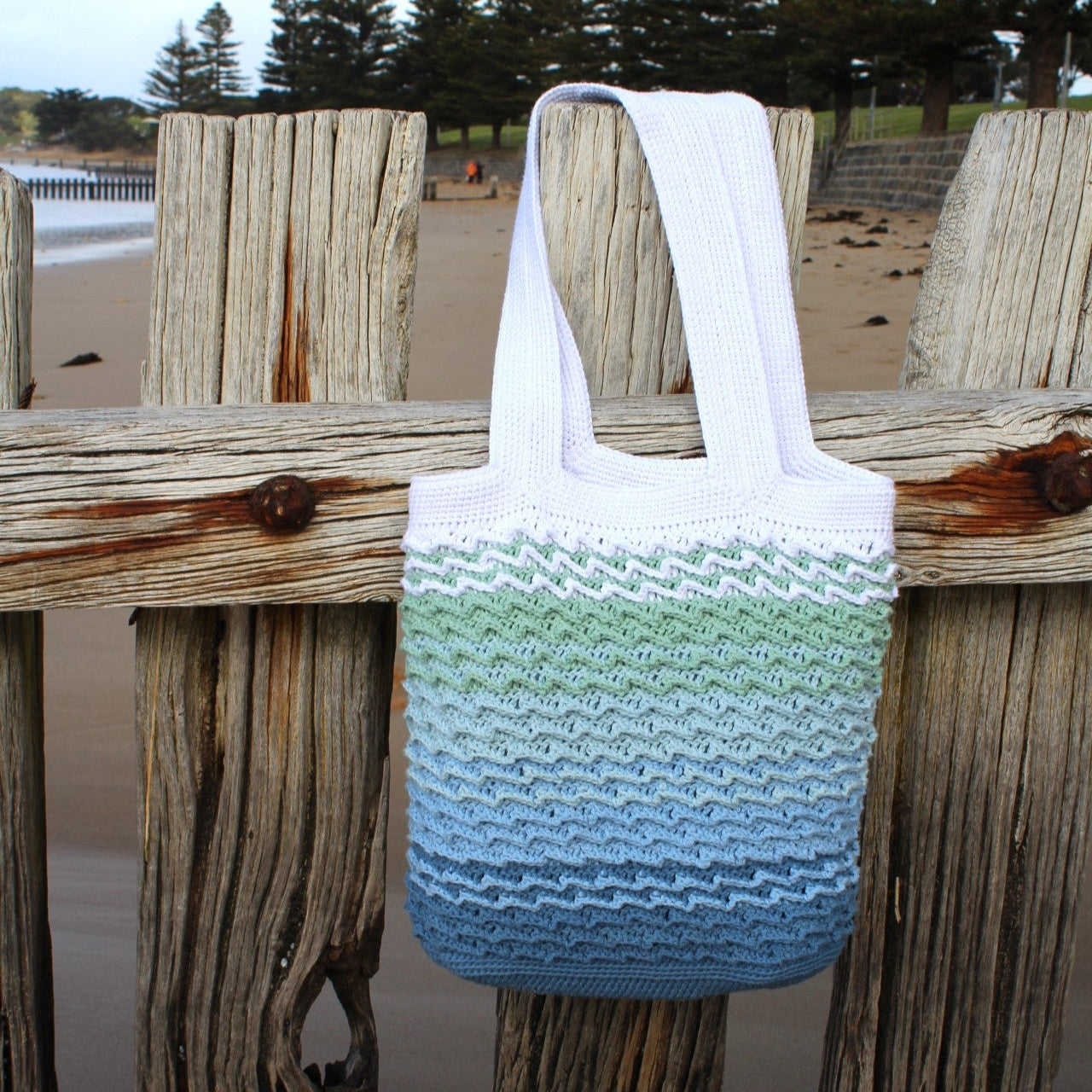 Knitted beach bag with gradient pattern on a wooden fence by a beach.