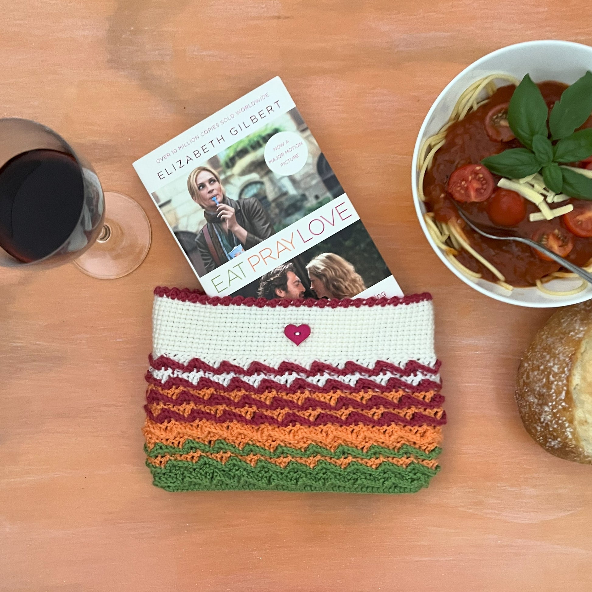 Colorful crocheted pouch on a table with a book, bowl of pasta, and glass of red wine.