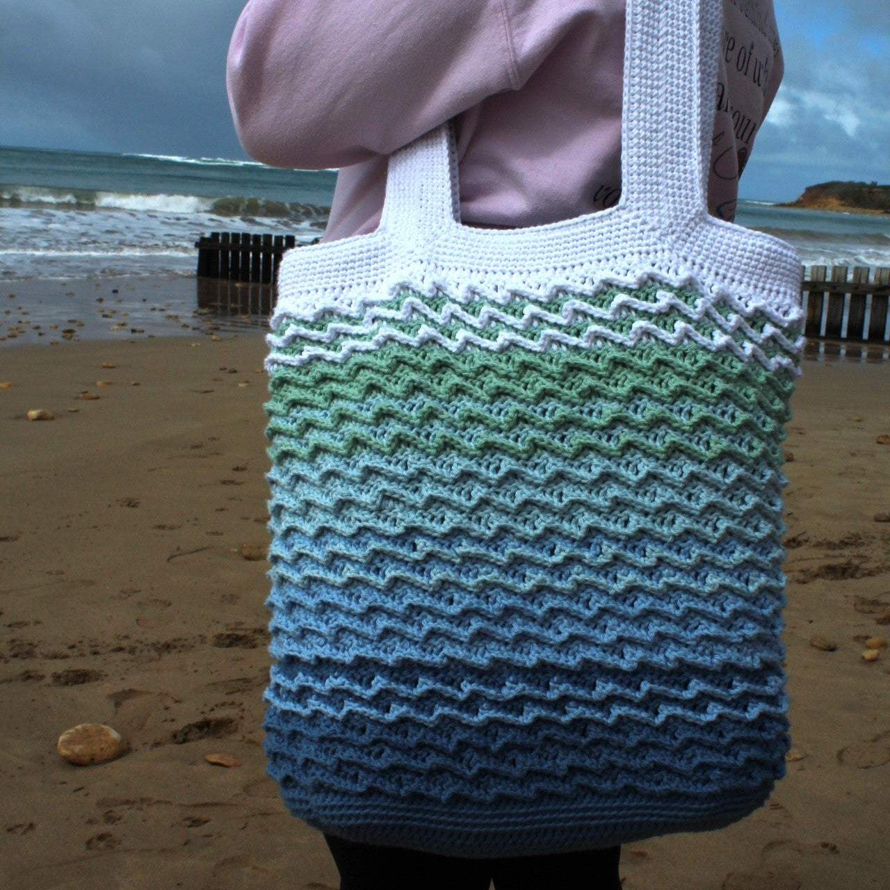 Person holding a large, colorful knitted bag on a beach.