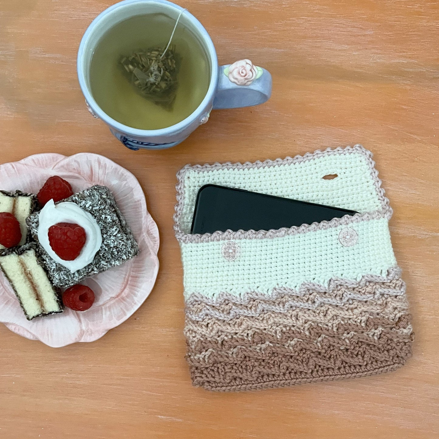 Cup of tea with a teabag, plate of snacks, and a crocheted pouch on a wooden surface.