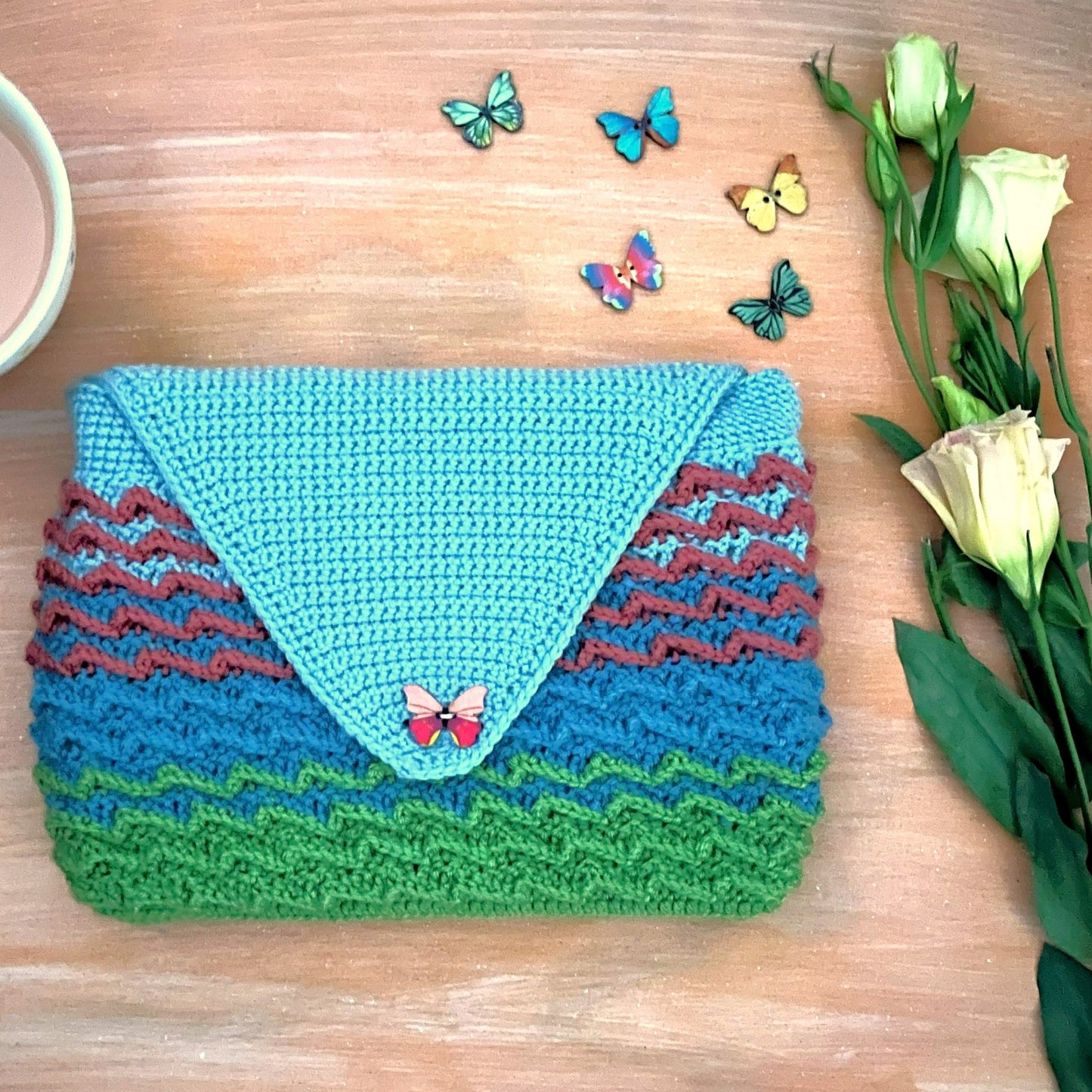 Crocheted pouch with butterfly design on a wooden surface with a mug and flowers.