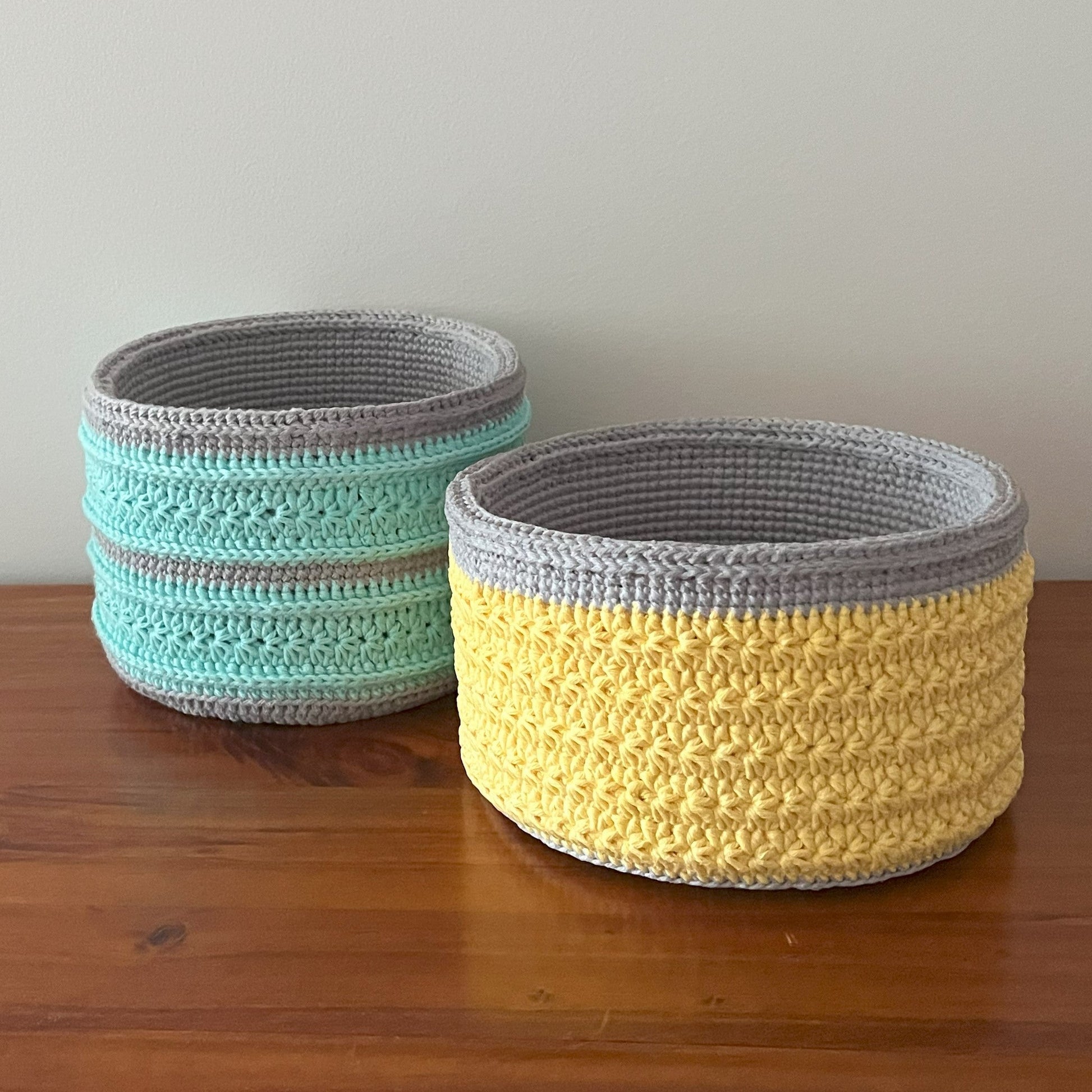 Two crocheted baskets, one green and one yellow, on a wooden surface with a plain background.