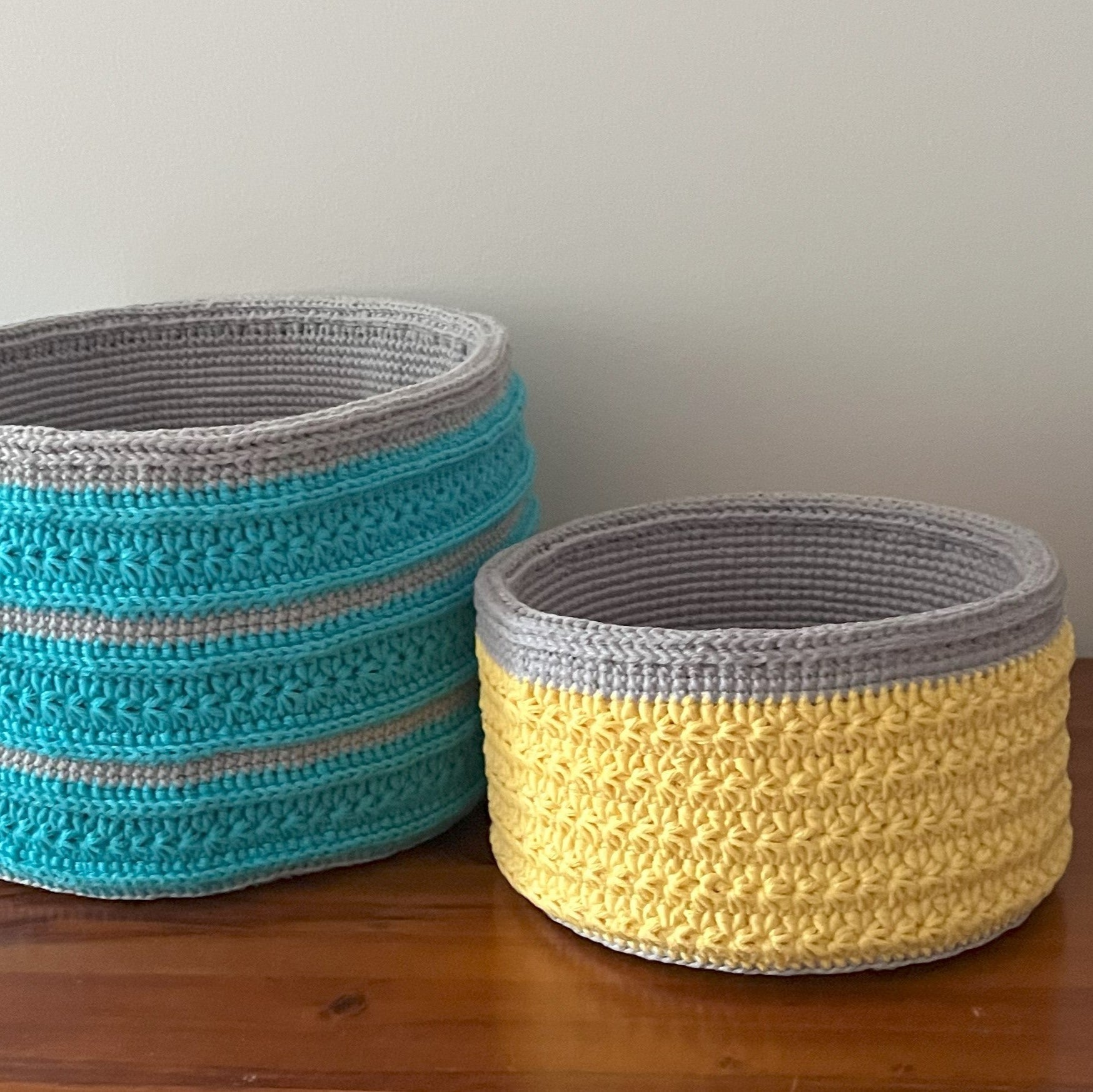 Two crocheted baskets, one blue and gray, the other yellow and gray, on a wooden surface.