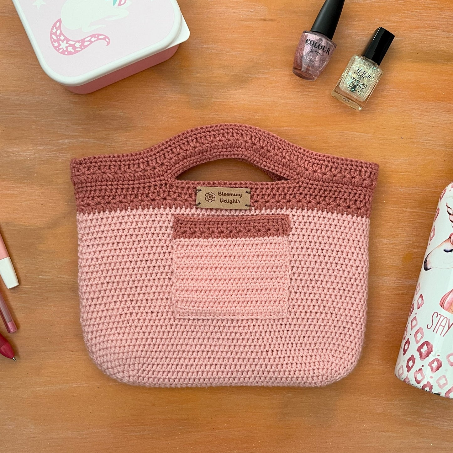 Pink crochet bag with a unicorn-themed water bottle and nail polish on a wooden surface