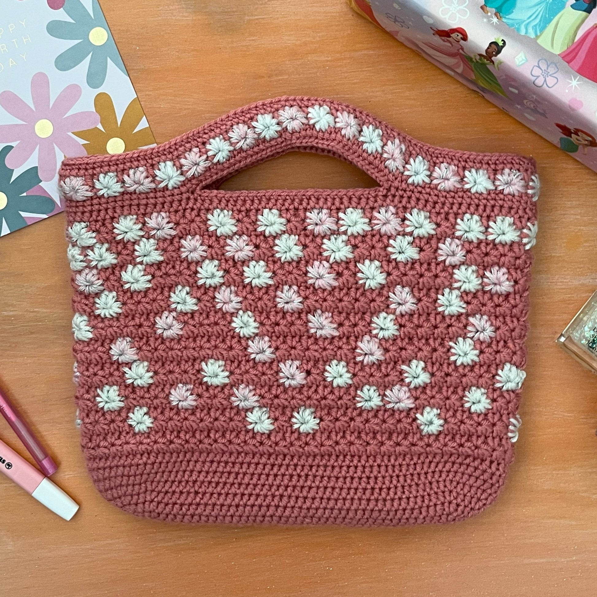 Pink crochet bag with white floral pattern on a wooden surface with stationery items.