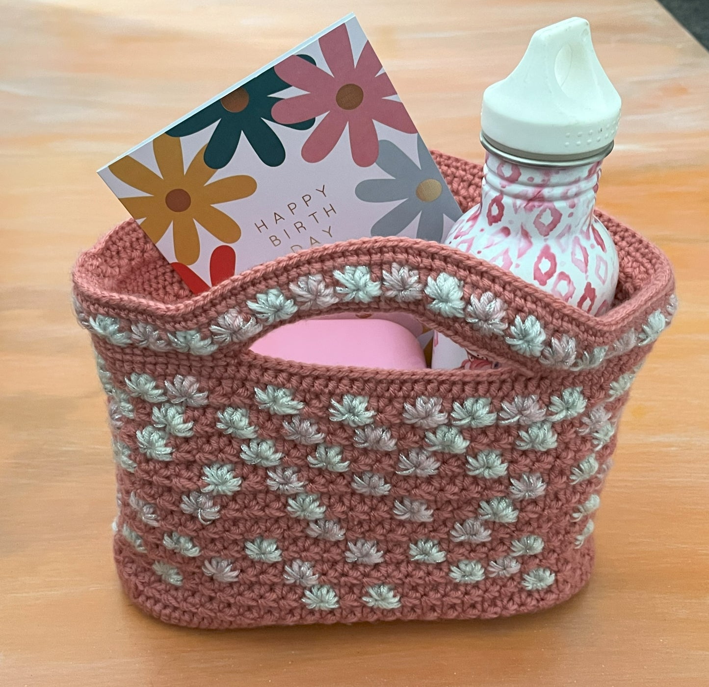 Crocheted basket with a bottle, card, and pink item on a wooden surface