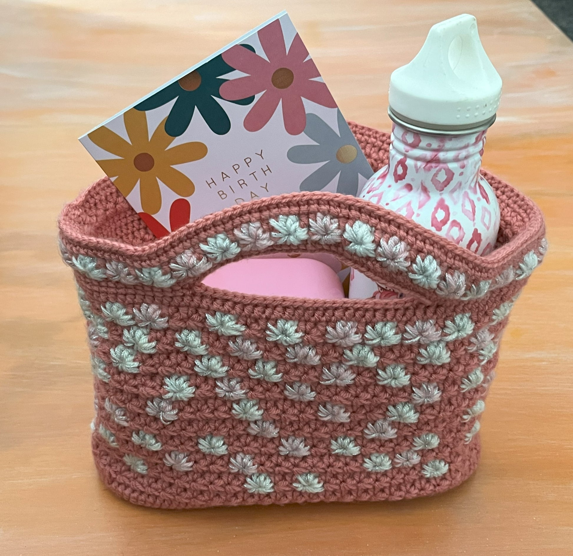 Crocheted basket with a bottle, card, and pink item on a wooden surface