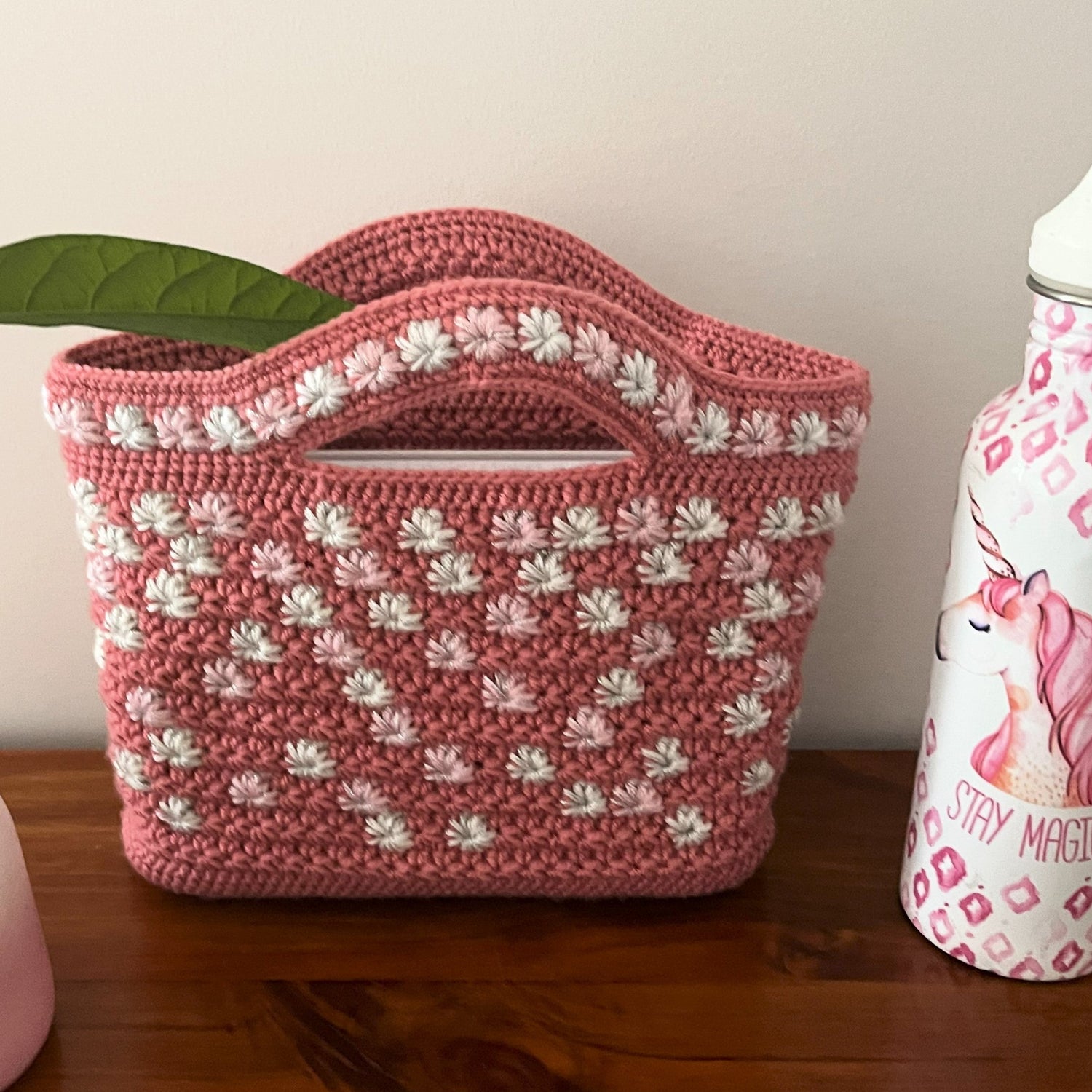 Pink crochet bag with white patterns on a wooden surface next to a pink bottle with a plant in the background.
