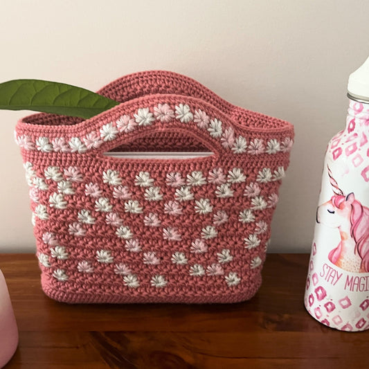 Pink crochet bag with white patterns on a wooden surface next to a pink bottle with a plant in the background.