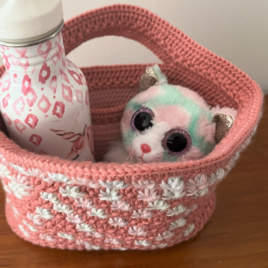 Pink and white crochet basket with a bottle and plush toy on a wooden surface