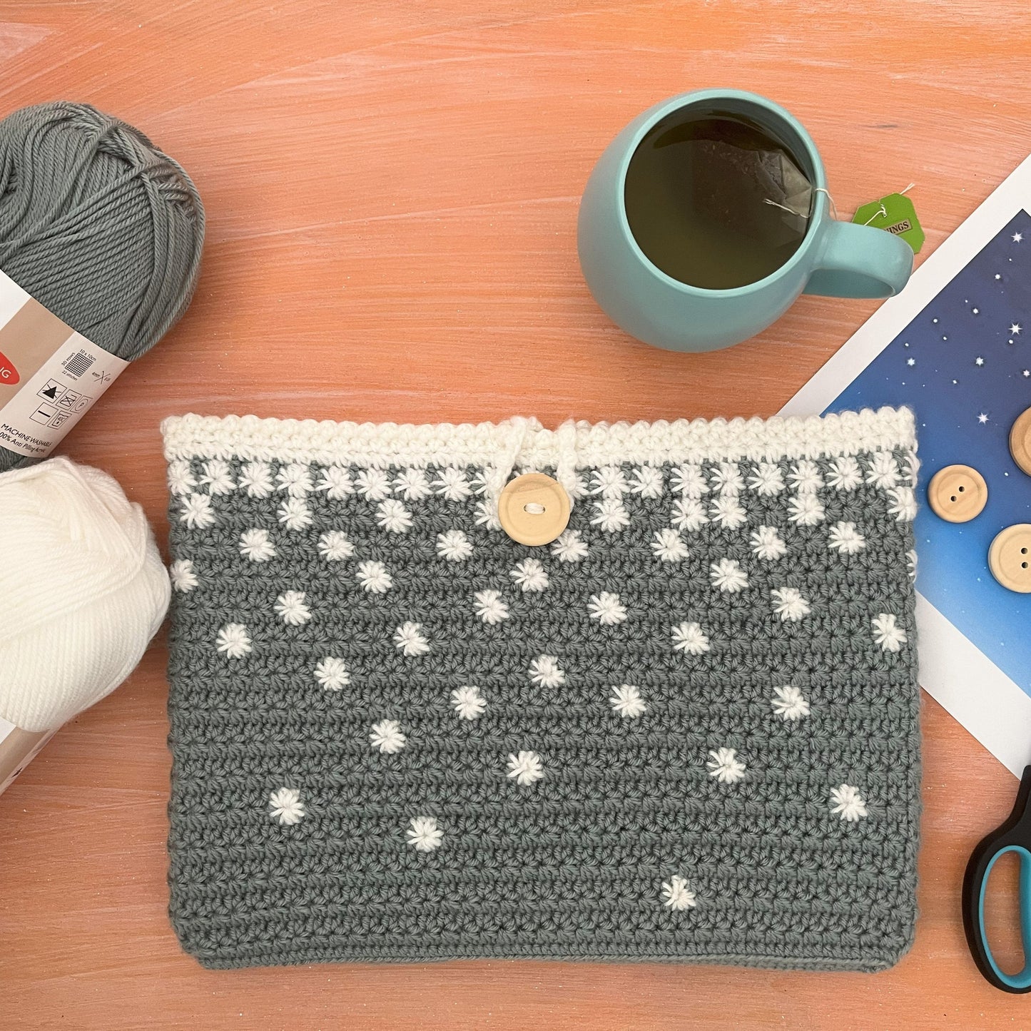 Crocheted pouch with white patterns on a wooden surface with yarn, mug, and scissors.