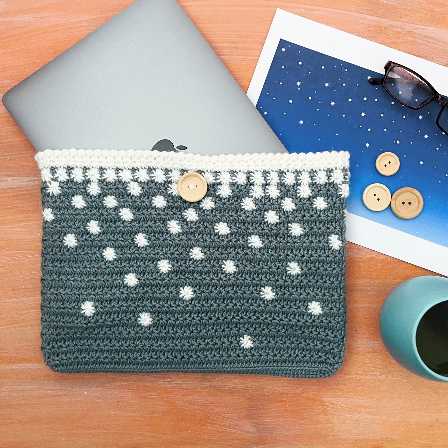 Crocheted pouch with a laptop on a wooden surface with a mug and photo album.