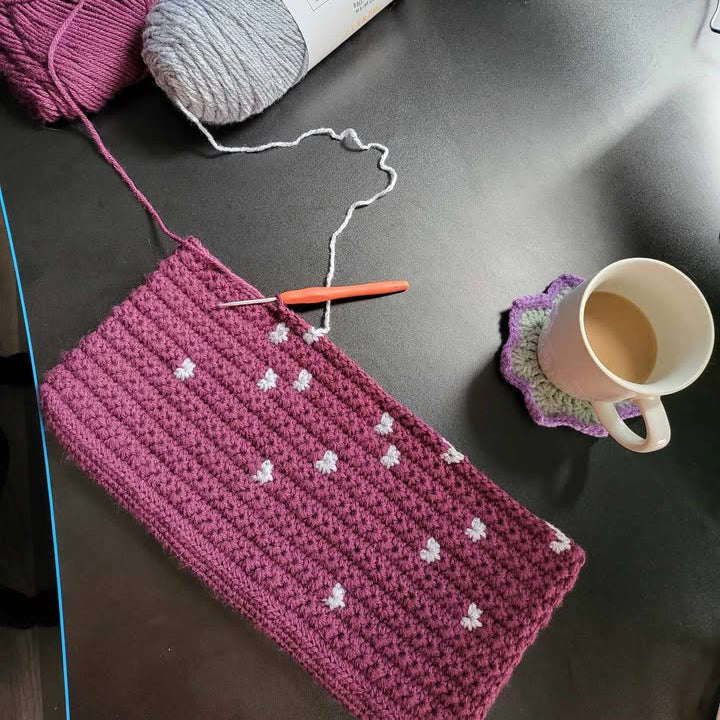 Pink crocheted item with white falling stars patterns on a dark surface, next to yarn and a mug.