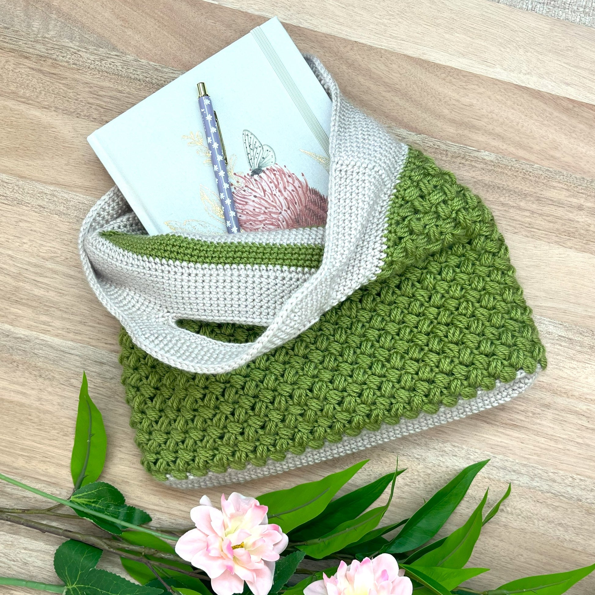Green and white crocheted bag with a notebook and pen on a wooden surface, surrounded by flowers and leaves.