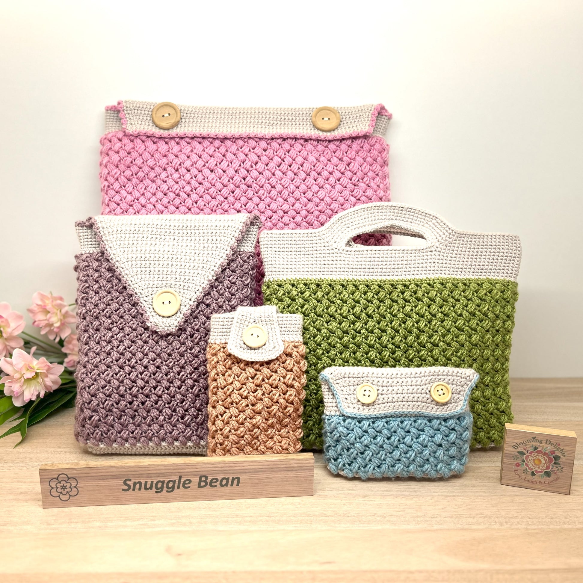 Set of crocheted bags in various colors with 'Snuggle Bean' branding on a wooden stand.