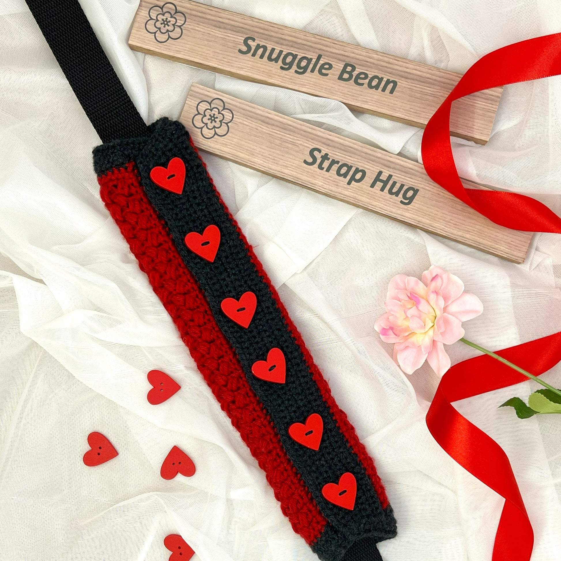 Red and black strap with heart patterns, wooden tags labeled 'Snuggle Bean' and 'Strap Hug', and decorative ribbons on a white background.