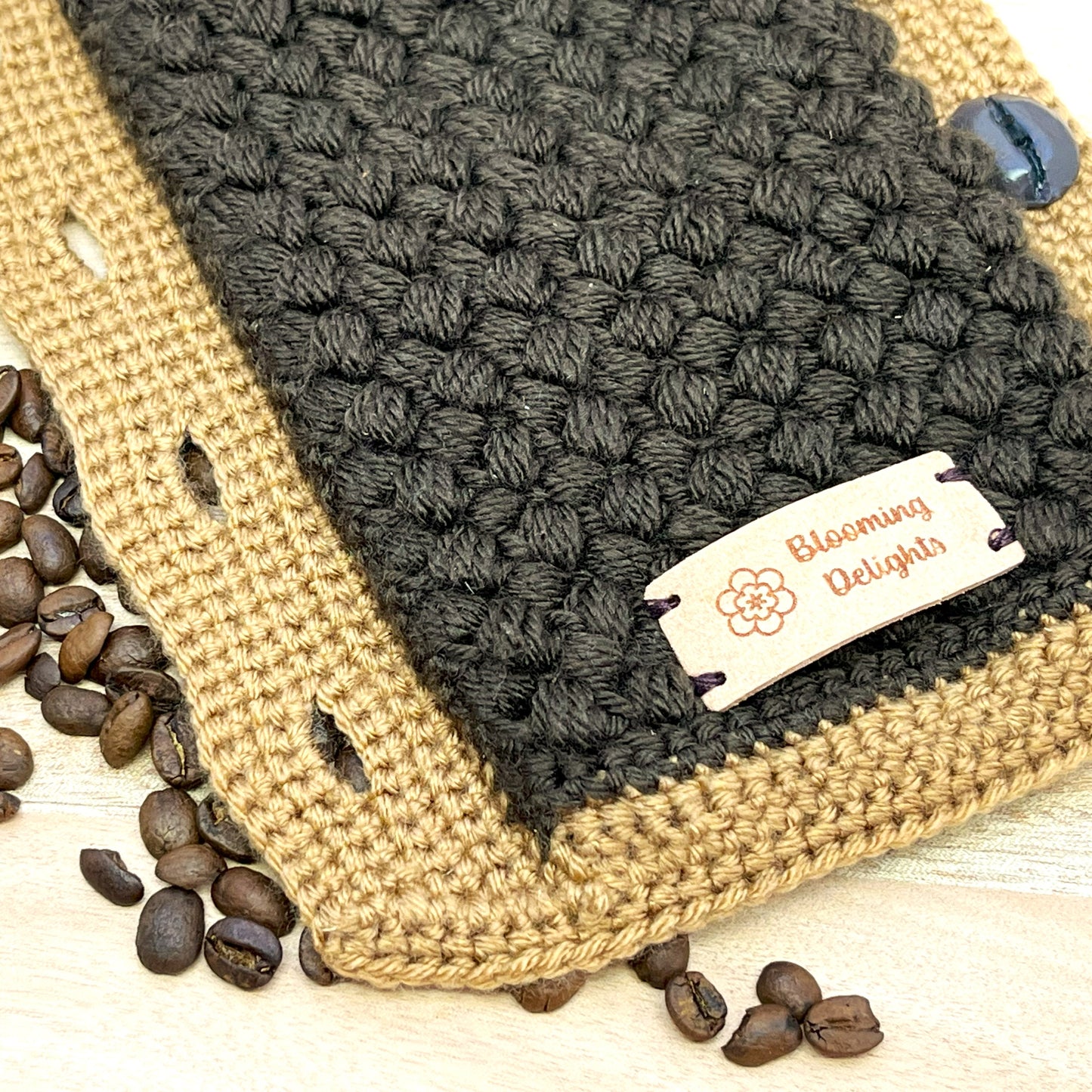 Handmade crochet coffee cozy with 'Blooming Delights' label on a wooden surface with coffee beans.