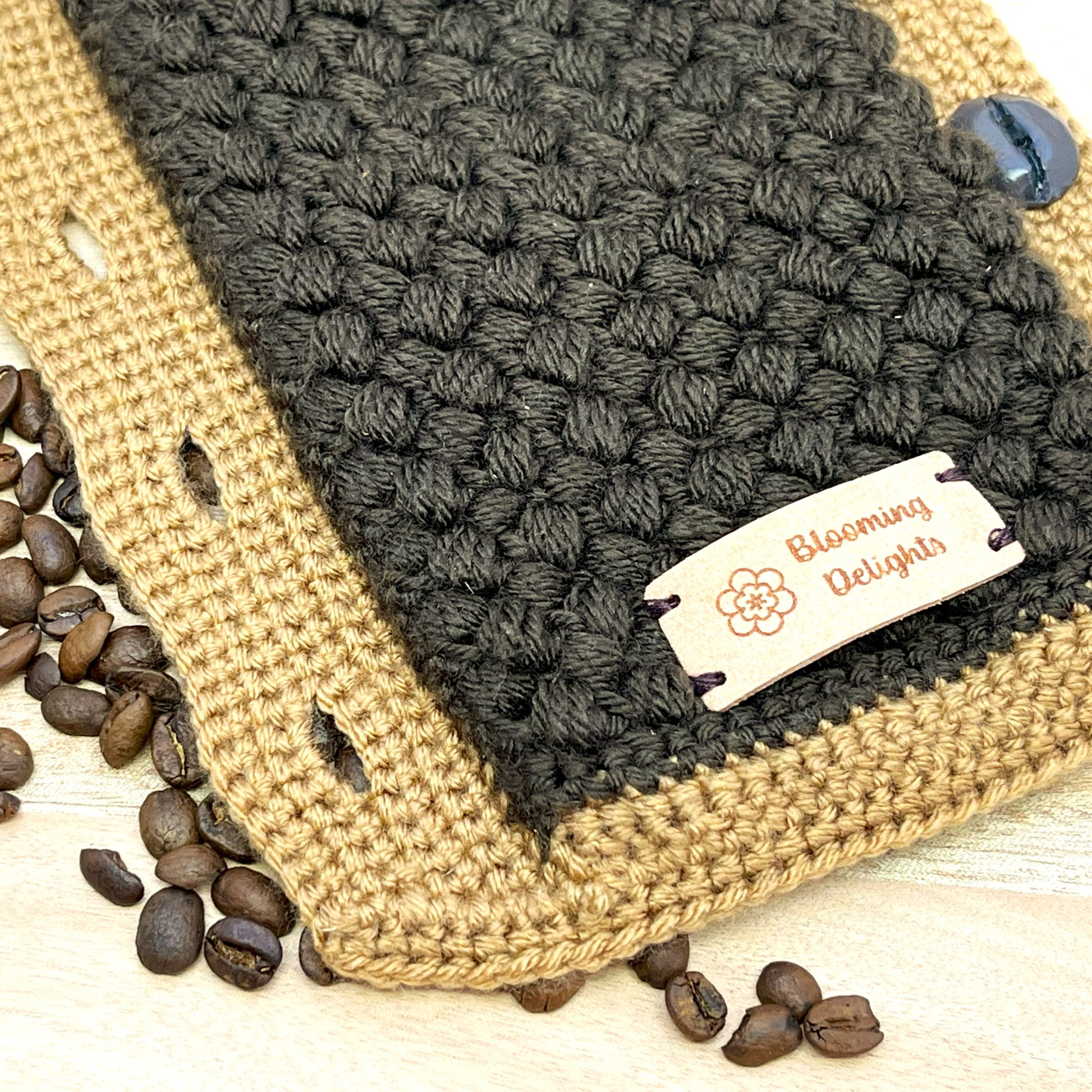 Handmade crochet coffee cozy with 'Blooming Delights' label on a wooden surface with coffee beans.
