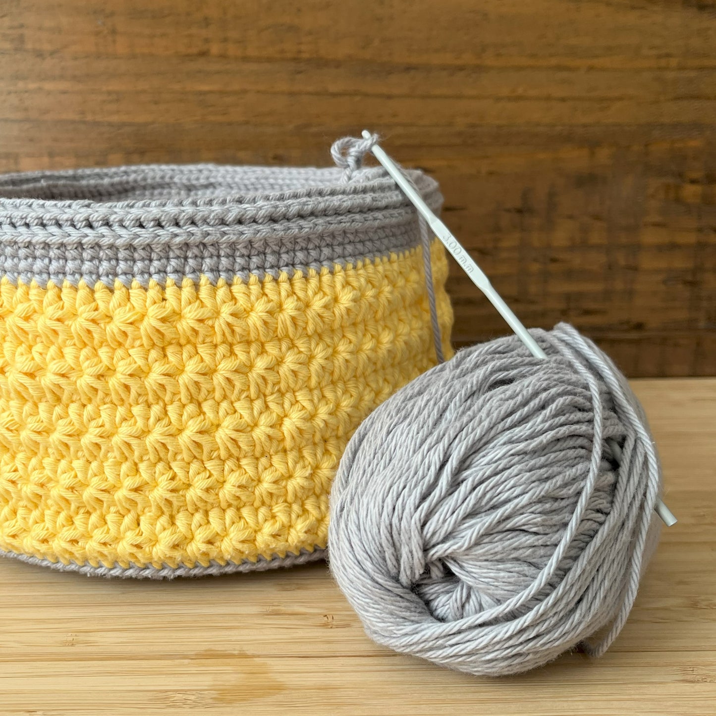 Crocheted basket with gray and yellow pattern and a ball of gray yarn on a wooden surface.