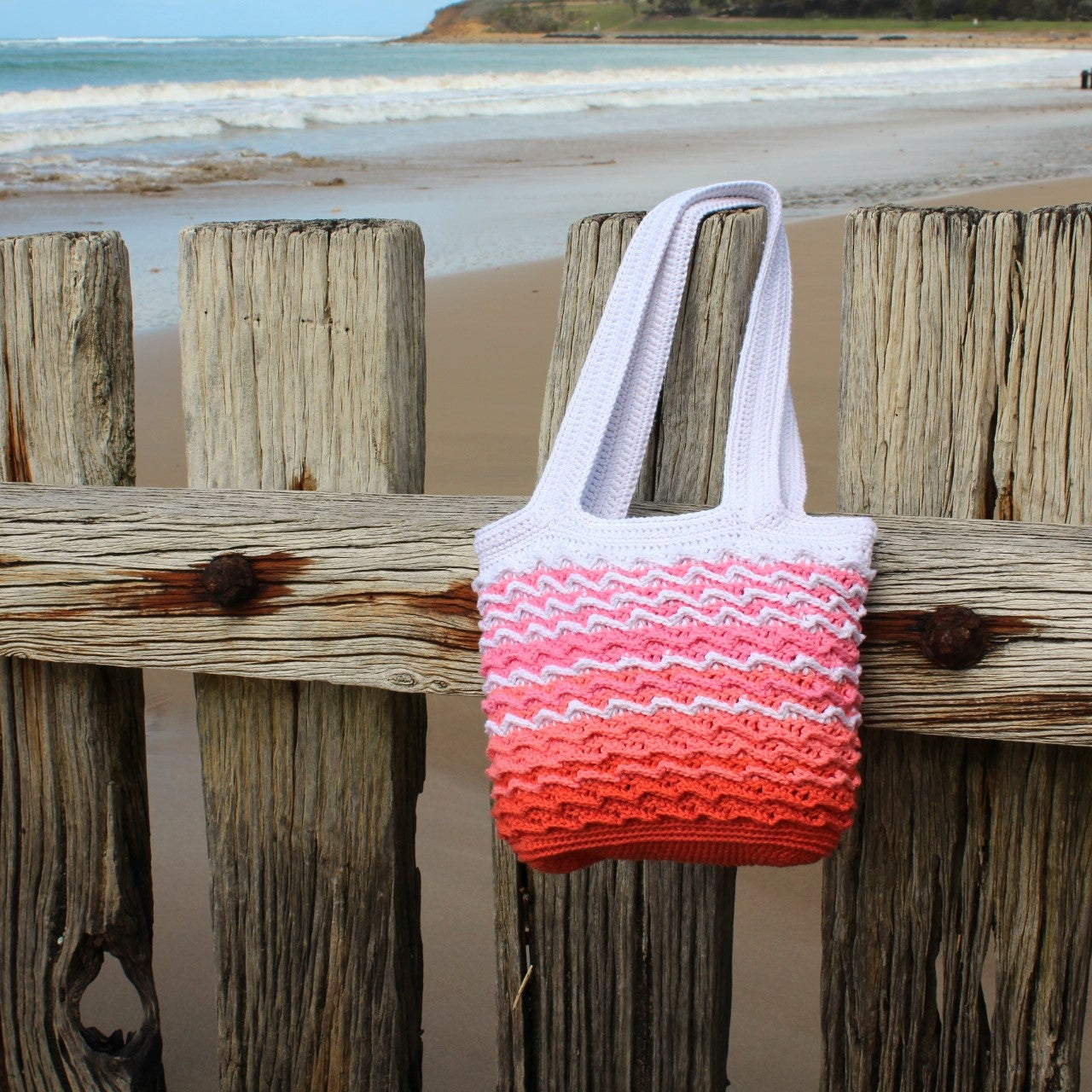 Crocheted bag with a gradient pattern on a wooden fence by a beach.